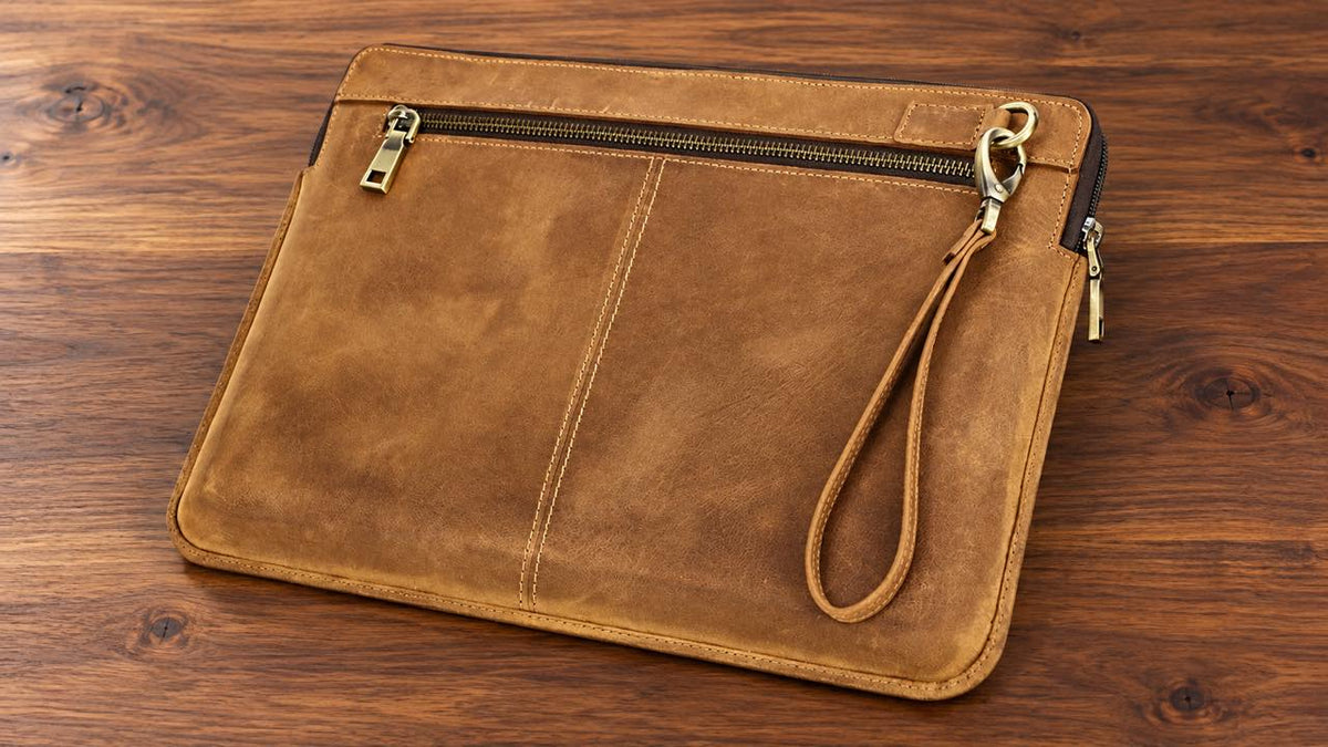 Tan leather MacBook sleeve with zipper and wrist strap on a walnut desk